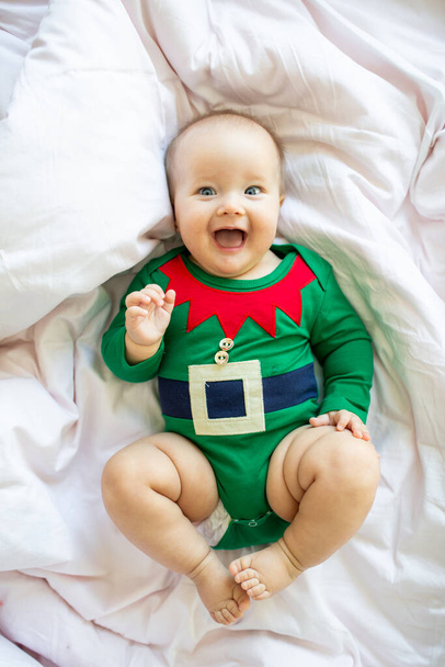 Cute caucasian baby girl six months old wearing elf costume green color. Adorable Santa helper lying on bed at home on white background - Fotoğraf, Görsel