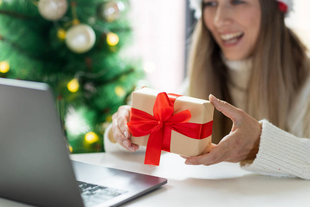 Young woman opening Christmas or new year present when calling to friends via video chat from home - Foto, Imagen