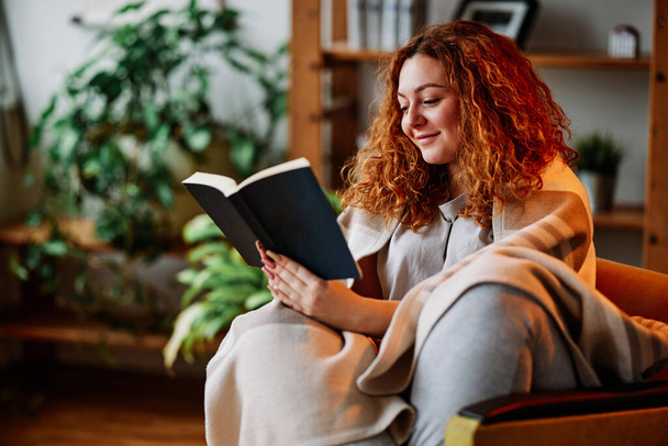 Рыжая наслаждается книгой дома. Счастливая рыжая девушка сидит в своем уютном доме и читает книгу. - Фото, изображение