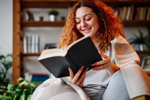 Una pelirroja leyendo un libro en casa. Una chica pelirroja feliz se sienta en un sillón cubierto con una manta y lee un libro interesante en su acogedora casa. - Foto, imagen