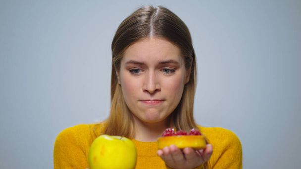 Portrait of thoughtful woman selecting between apple or cake at camera. - Photo, Image