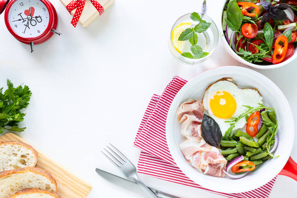 Frühstück für die Liebsten im Urlaub: herzförmiges Ei, Speck, grüne Bohnen auf weißem Tisch. Selektiver Fokus. Blick von oben. Kopierraum - Foto, Bild