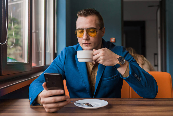 Ein junger, gutaussehender Geschäftsmann von europäischem Aussehen sitzt mit Smartphone oder Handy in einem Café an einem Tisch in einer Kaffeepause. - Foto, Bild