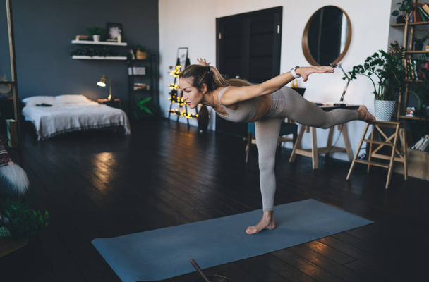 Europejska dziewczyna ćwiczy jogę na macie fitness w domu. Młoda, skoncentrowana kobieta w sportowej odzieży stojąca w pozycji wojownika i balansująca na jednej nodze z zamkniętymi oczami. - Zdjęcie, obraz