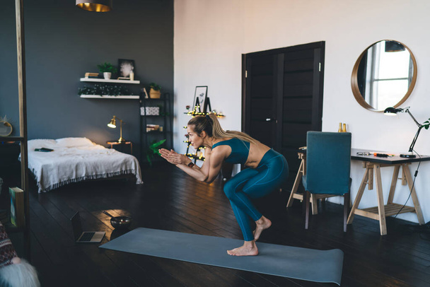 European girl practicing yoga on fitness mat at home. Concept of healthy lifestyle. Young beautiful focused athletic woman in sportswear and barefoot. Interior of studio apartment at daytime - 写真・画像