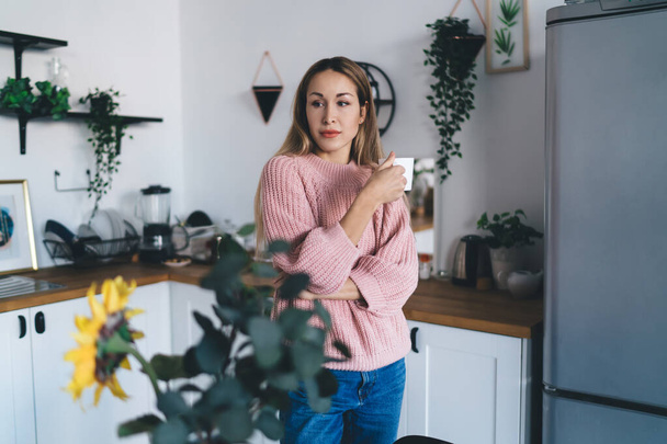 Thoughtful european girl drinking tea from cup at home. Concept of domestic lifestyle. Idea of home rest and leisure. Young beautiful woman wear sweater and jeans. Interior of kitchen in modern flat - Фото, изображение