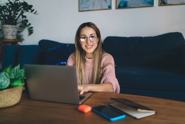 European girl in glasses using laptop computer at home. Concept of domestic lifestyle. Idea of home rest and leisure. Young beautiful smiling woman sitting at table. Interior of studio apartment - 写真・画像
