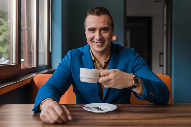 Elegante, adulto atractivo hombre de apariencia europea en un traje se sienta en una mesa en un café y bebe café en una pausa para el almuerzo. - Foto, Imagen