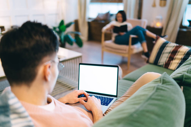 Man using laptop on sofa while her blurred girlfriend reading book in armchair. young couple resting at home. Concept of domestic entertainment and leisure. Interior of modern spacious apartment - Photo, Image