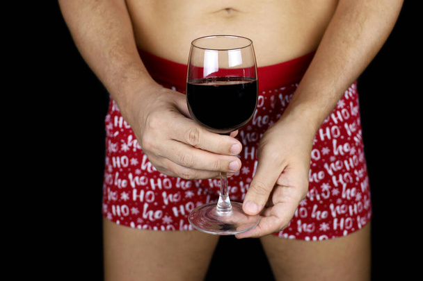 Man in with naked torso wearing Christmas underpants standing with glass of red wine in hands on black background. Concept of New year celebration, alcohol abuse, romantic sex date - Photo, image