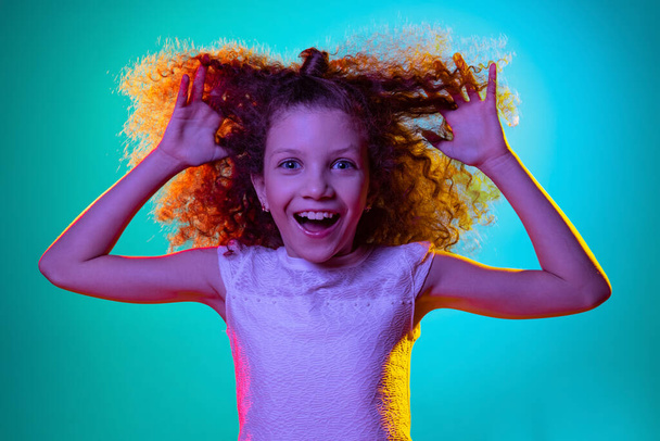 Close-up little cute curly preschool beautiful girl looking at camera isolated on blue studio background in purple neon light. - Foto, imagen