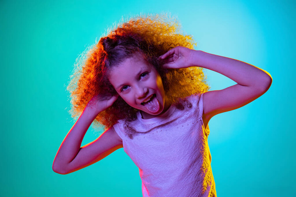 Half-length portrait of cute curly preschool beautiful girl with red hair making faces isolated on blue studio background in purple neon light. - Foto, afbeelding