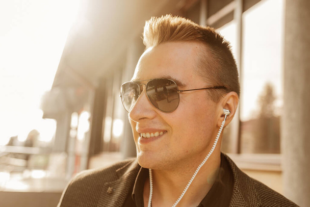 Porträt eines positiv lächelnden jungen Geschäftsmannes europäischen Aussehens mit Sonnenbrille und Kopfhörer, der draußen auf der Straße Musik hört. - Foto, Bild