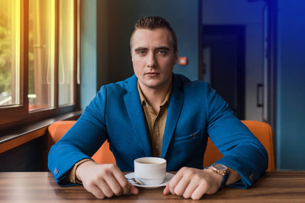 青いジャケットと茶色のシャツの中の白人の外観の収集された大人、ビジネスマンの肖像画は休憩中にコーヒーを飲みながらカフェのテーブルに座っています. - 写真・画像