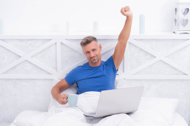 Happy man drink coffee and use laptop stretching after sleep in bed in morning, wake up - Φωτογραφία, εικόνα