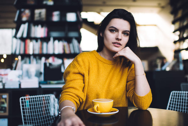 Thoughtful young female in trendy sweater leaning on elbow and touching chin while resting at table with cup of coffee in contemporary cafe - Foto, Imagem