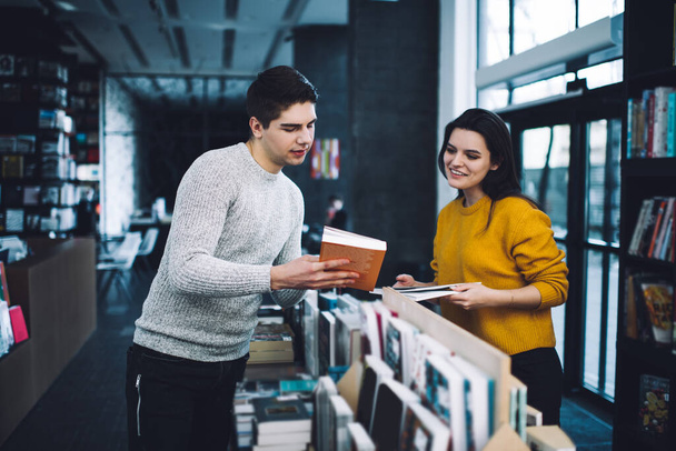 Positive couple choosing books together among stands with presented products while man showing jacket of book to smiling interested woman in store - 写真・画像
