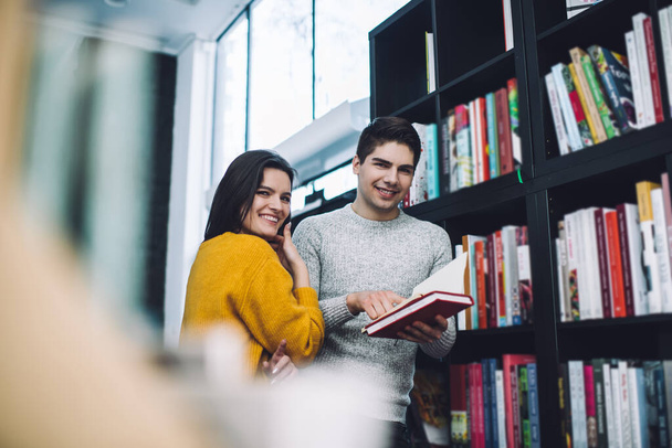 From below of young cheerful man and woman in warm outfits standing near bookshelves  with opened textbook and looking at camera - Fotoğraf, Görsel