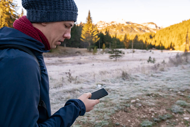 Un joven en una bufanda y un sombrero está buscando algo en el teléfono contra el fondo de una montaña y hierba congelada de la mañana - Foto, Imagen
