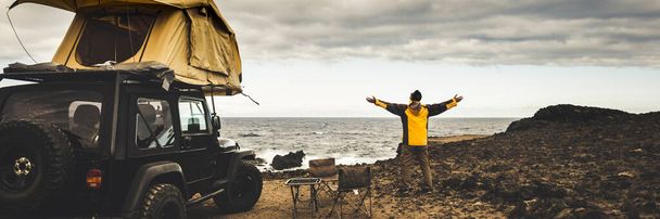 Hombre abriendo brazos y disfrutando de la naturaleza en la actividad de vacaciones de aventura salvaje. Coche con tienda en el techo y vista al mar - Foto, imagen