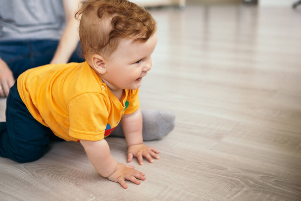 Ragazzino sorridente allegro carino bambino rossa ridendo bambino imparare a strisciare giocando sul pavimento. Bambino divertirsi a casa vestito arancione t-shirt. Infanzia felice, concetto di famiglia - Foto, immagini