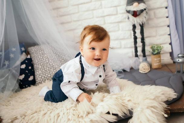 Mały chłopiec uśmiechnięty wesoło słodkie dziecko rudowłose śmiejące się dziecko uczące się pełzania bawiące się na podłodze dywan. Maluch bawiący się w domu ubrany w szelki i białą koszulę. Szczęśliwego dzieciństwa - Zdjęcie, obraz