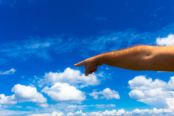 Fotografia męskiej ręki skierowanej w jednym kierunku, na tle błękitnego nieba z białymi letnimi chmurami; przestrzeń kopiująca - Zdjęcie, obraz