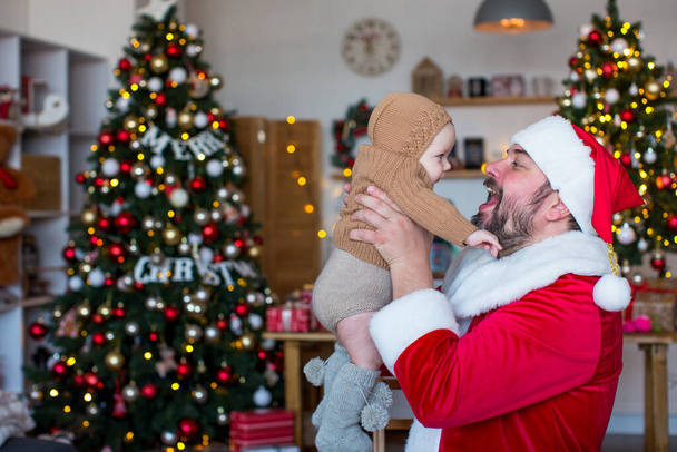 Noel Baba, Noel ağaçlarının arka planında onu kollarında tutan bir çocukla oynuyor. - Fotoğraf, Görsel