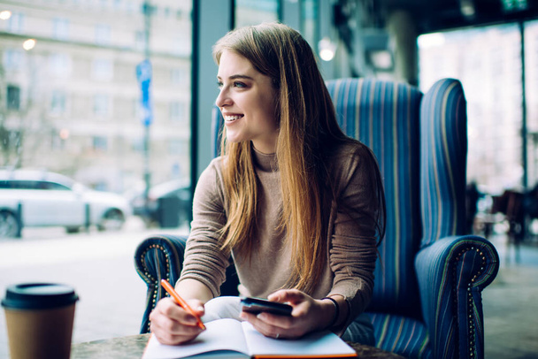 Smiling female freelancer in casual wear sitting in comfortable armchair and messaging on smartphone while taking notes and looking away - Foto, afbeelding