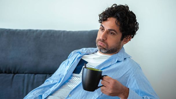 Handsome male model feels tired after work and having a cafe. Mature man drinking morning expresso coffee while having headache while sitting on couch home. Unpleasant pain. - Photo, image