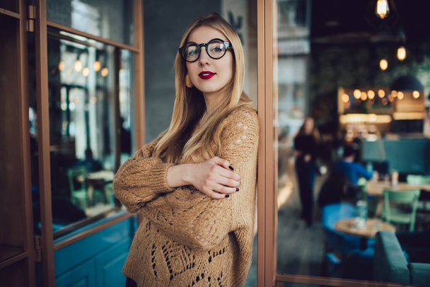 Thoughtful female in casual sweater and glasses standing in modern cafe and looking at camera while spending time in afternoon - Zdjęcie, obraz
