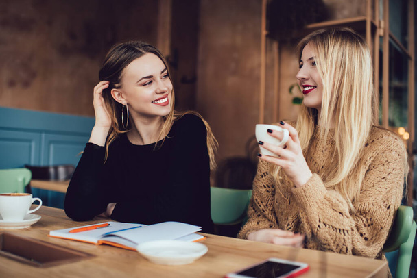 Smiling female friends in casual clothes drinking cup of hot coffee while sitting together at wooden table in modern cafe and looking at each other - Foto, immagini