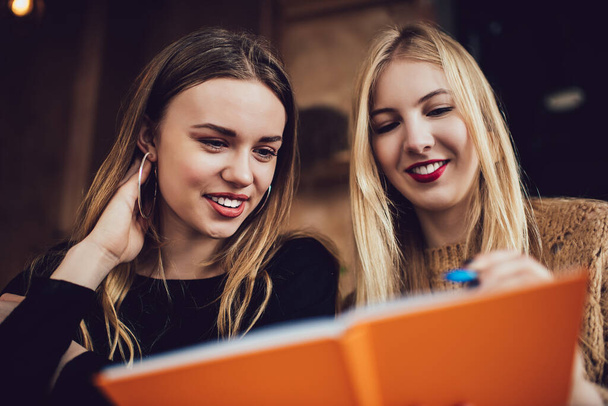 Cheerful female friends in casual clothes sitting at wooden table and writing ideas in copybook while enjoying time together in cafe - Foto, afbeelding