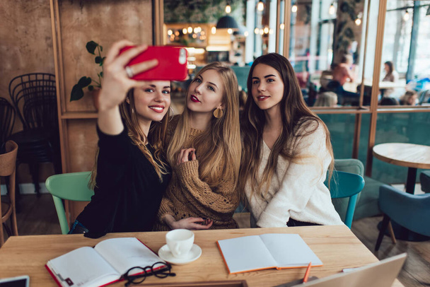 Charming young female friends in casual clothing with long hair taking selfie and looking at camera of smartphone with smiles while doing homework together in time cafe - Valokuva, kuva
