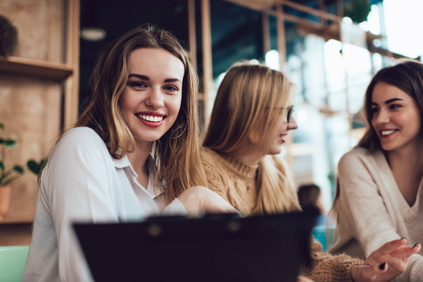 Positive woman in casual wear smiling and looking at camera while sitting in modern cafe with clipboard during work with friends - Zdjęcie, obraz