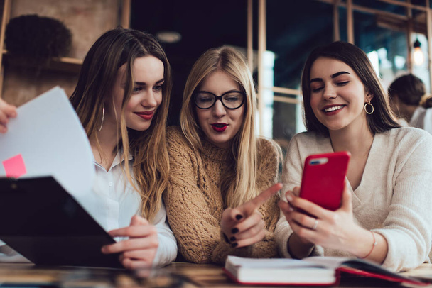 Low angle of group of joyful women in casual wear sitting together in modern cafe and browsing smartphone while working with clipboard - Фото, изображение