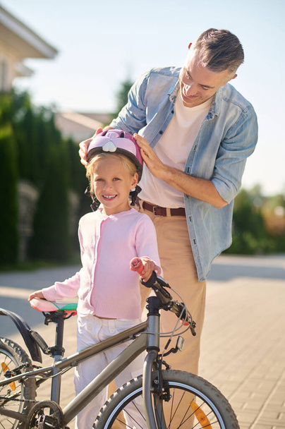 A man putting protective helmet on his daughters head before riding a bike - Foto, Imagen