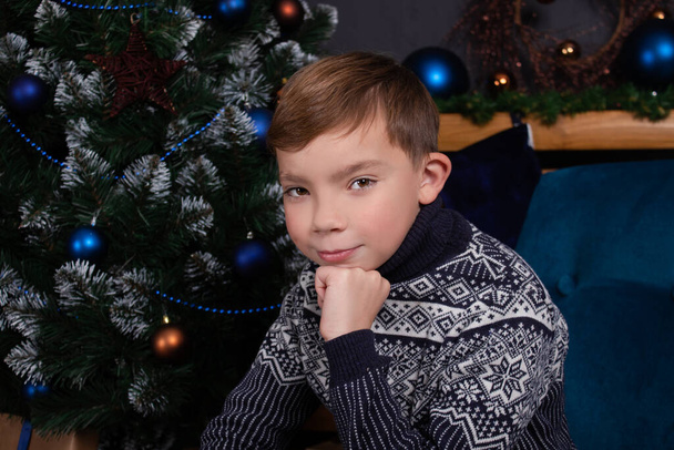 charming blonde teenage boy in a knitted dark blue sweater near decorated christmas tree. happy holidays. new years eve - Foto, afbeelding
