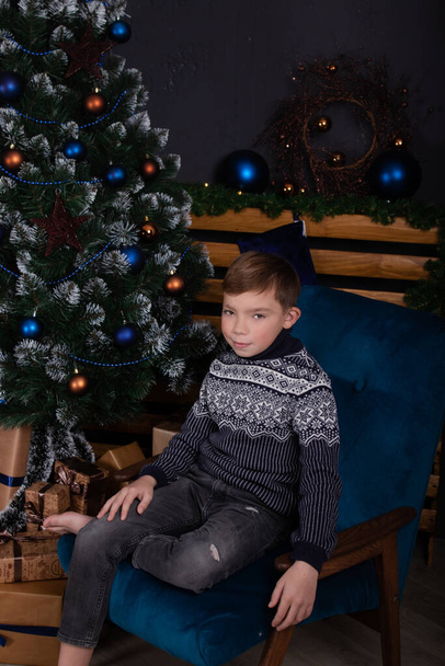 bájos szőke tinédzser fiú egy kötött sötétkék pulóverben a díszített karácsonyfa mellett. Kellemes ünnepeket. szilveszterkor - Fotó, kép