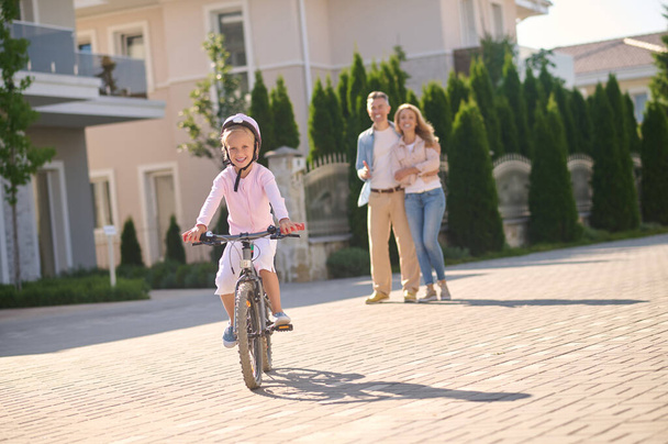 Ένα κορίτσι να κάνει ποδήλατο, ενώ οι γονείς της να την παρακολουθούν. - Φωτογραφία, εικόνα