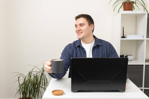 Ένας άντρας δουλεύει σε ένα λάπτοπ ενώ κάθεται σε ένα τραπέζι στο σπίτι. Η έννοια της απομακρυσμένης εργασίας κατά τη διάρκεια καραντίνας. Ο Freelancer εργάζεται στον υπολογιστή. - Φωτογραφία, εικόνα