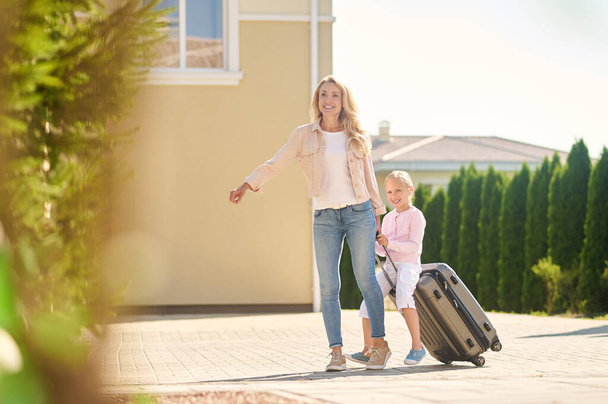Blonde woman carrying a suitcase with her daughter riding it - Foto, Bild