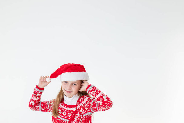 幸せな女の子のダンスの肖像画,白い背景に隔離された赤いクリスマスセーターとサンタの帽子を身に着けています。若いですcaucasian喜び笑顔ブロンド女の子でAクリスマス赤い鹿のセーターは幸せです. - 写真・画像