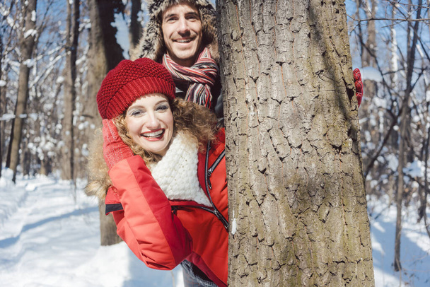 Couple ludique se cachant derrière un tronc d'arbre dans la neige - Photo, image