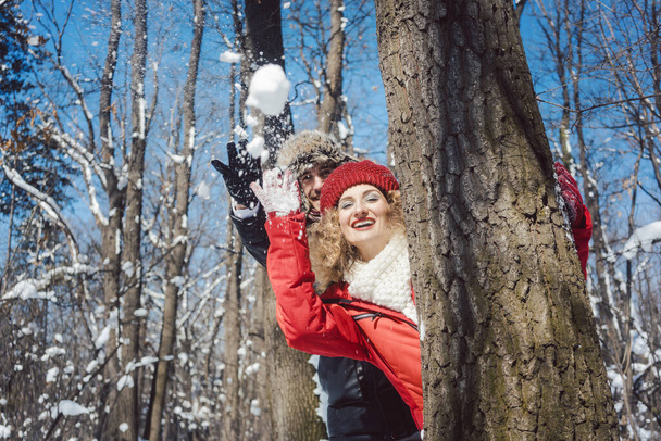 Woman and man in winter throwing snowball hiding behind a tree - Photo, Image