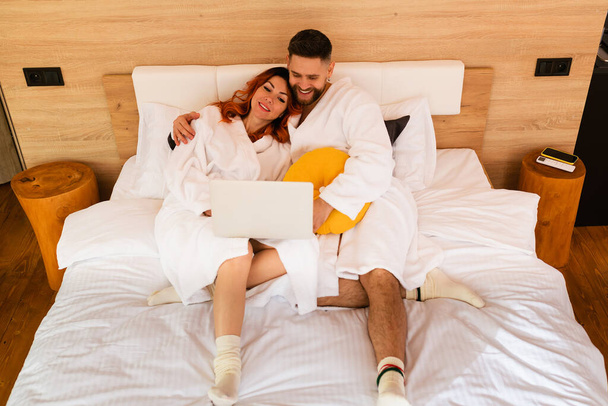 Άποψη πλήρους μήκους ενός νεαρού ζευγαριού με μπουρνούζια ξαπλωμένο στο κρεβάτι με φορητό υπολογιστή. Άντρας και γυναίκα βλέπουν ταινία. Άποψη από ψηλά. - Φωτογραφία, εικόνα