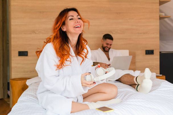 Kobieta ze słuchawkami śmieje się siedząc na łóżku ze skrzyżowanymi nogami. Rudowłosa kobieta w szlafroku ze słuchawkami. - Zdjęcie, obraz