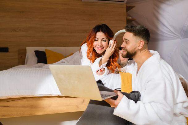 Foto de una joven pareja con albornoces acostada en la cama con un ordenador portátil. Hombre y mujer escuchan música. - Foto, imagen