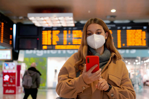 Traveler γυναίκα φορώντας FFP2 KN95 μάσκα προσώπου στο σιδηροδρομικό σταθμό. Γυναίκα μετακινούμενος ελέγχει το smartphone της με πίσω χρονοδιαγράμματα αναχωρήσεων αφίξεων. - Φωτογραφία, εικόνα