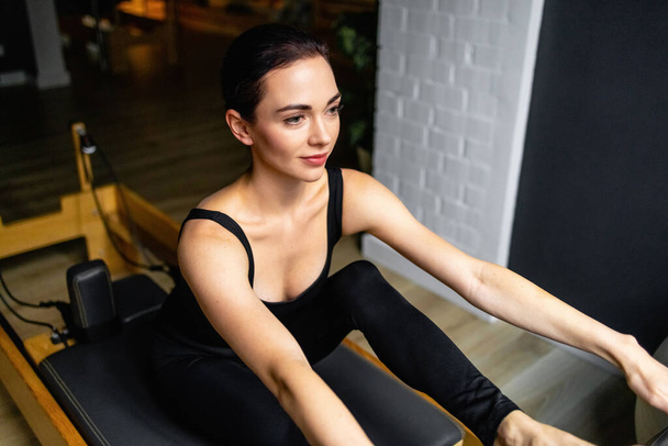 Sporty woman doing exercises on pilates reformer training machine. Working out wearing sportswear, gym. - Photo, Image